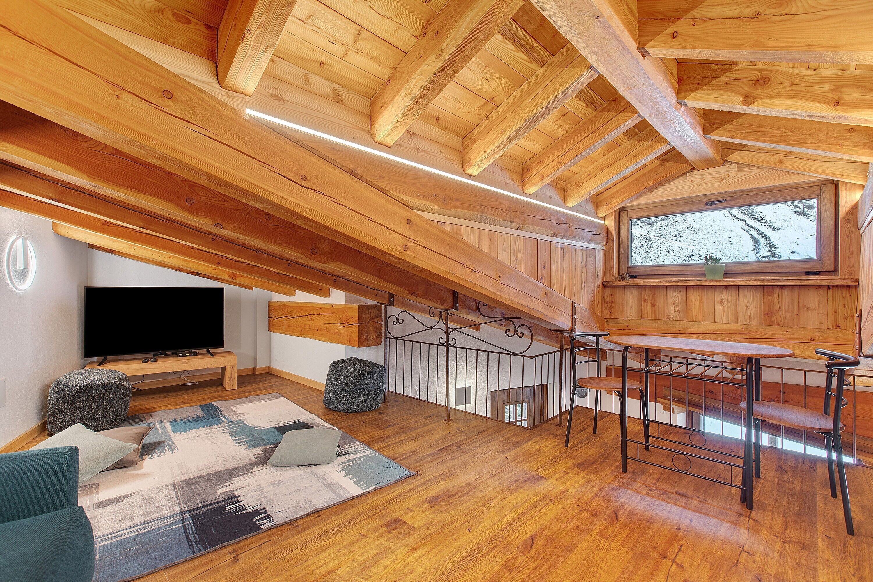 A loft-style room with a wooden ceiling, a television, a sofa, a coffee table, and a window with a view of trees.