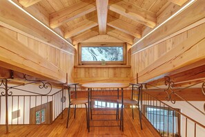 A wooden interior with a central table and chairs, a window with a view of trees, and a staircase with metal railings.