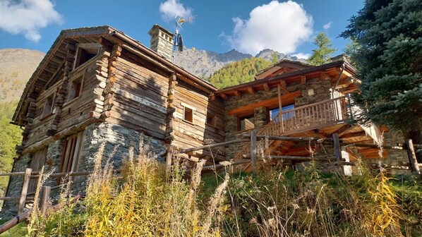 A rustic log cabin with a stone foundation, a balcony, and a mountain backdrop.