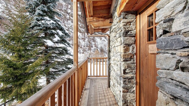 A wooden balcony with stone walls, a wooden railing, and a view of snow-covered trees.