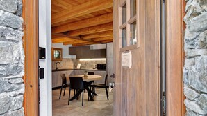 Eine Holztür führt in einen Essbereich mit einem runden Tisch und Stühlen. Im Hintergrund ist eine Küche erkennbar, und an einer Seite befindet sich eine Steinwand.