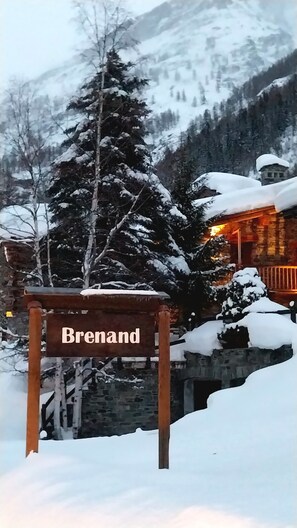 En träskylt med ordet ”Brenand” skrivet på den, en byggnad av sten med snö på taket och ett snötäckt berg i bakgrunden.