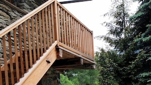 Ein Steinhaus mit einer Holztreppe, die zu einem Balkon führt, umgeben von Kübeln mit roten Blumen und einer Rasenfläche.