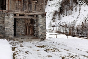 A rustic stone building with a wooden door, a stone staircase, and a snow-covered landscape with trees.