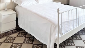 Una cama de metal blanco con cabecera decorativa, una mesita de noche blanca con lámpara, y un armario en una habitación con baldosas de diseño.