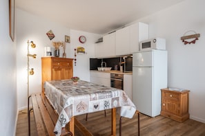 Una cocina compacta con armarios blancos, microondas y refrigerador. Incluye un banco de madera y una mesa con mantel estampado.
