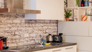 Une cuisine avec des armoires blanches, un mur en briques, une hotte de cuisinière en acier inoxydable, un évier, une cuisinière et un petit coin repas avec une chaise et une table.