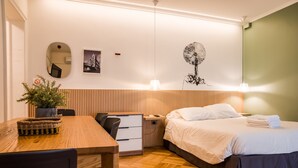 Habitación de hotel con piso de madera, cama con ropa blanca, escritorio con silla, una mesita con una planta y un cuadro con una representación de un árbol.