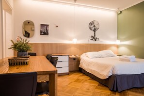 A hotel room with a wooden floor, a bed with white bedding, a desk with a chair, a small table with a plant, and a wall with a picture and a wall art of a tree. - Studio Oasis | Near Bellavista and Hospitals (Providencia)