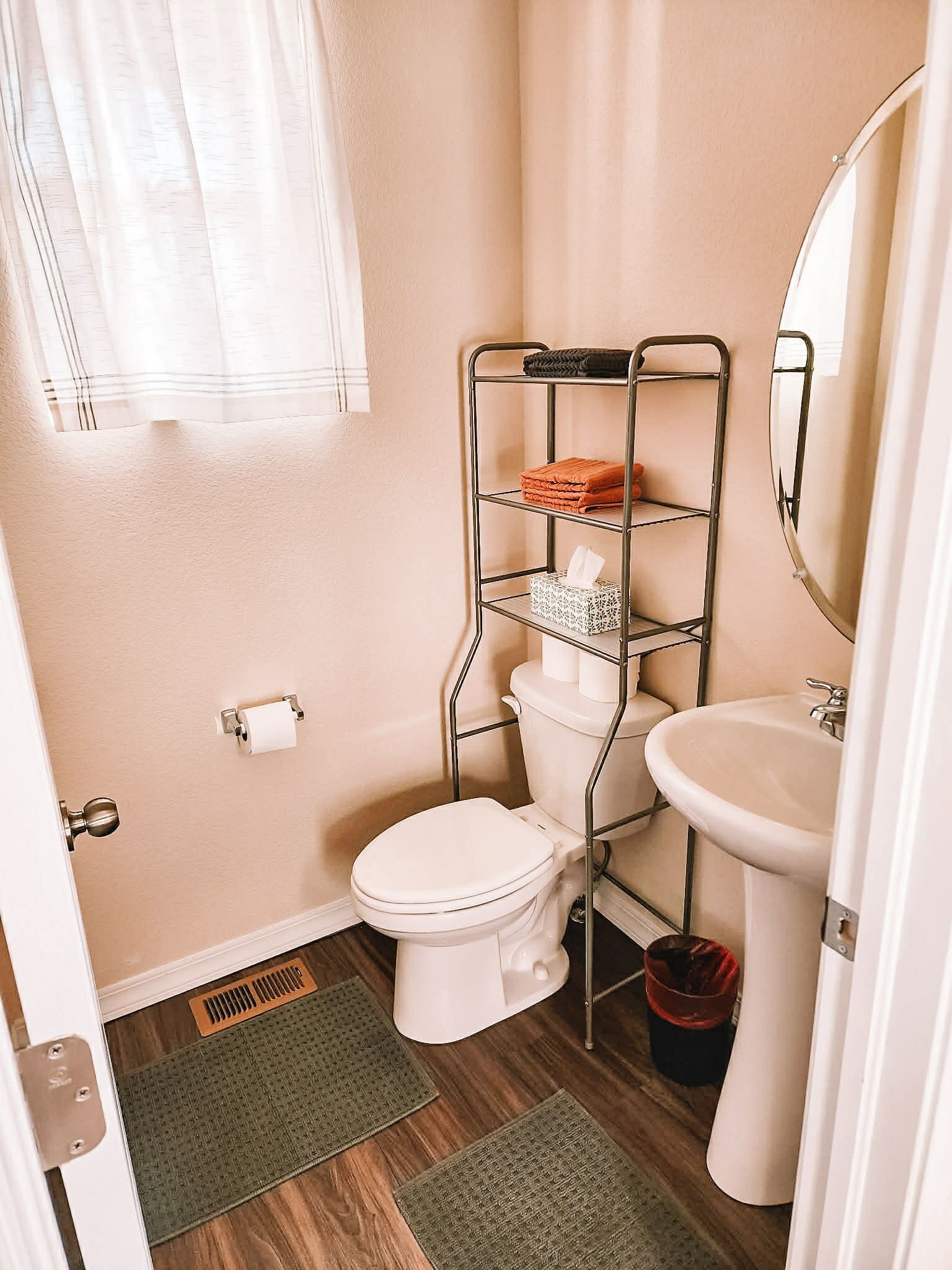 A compact bathroom with a toilet, a metal shelf unit, a sink, and a window with curtains.