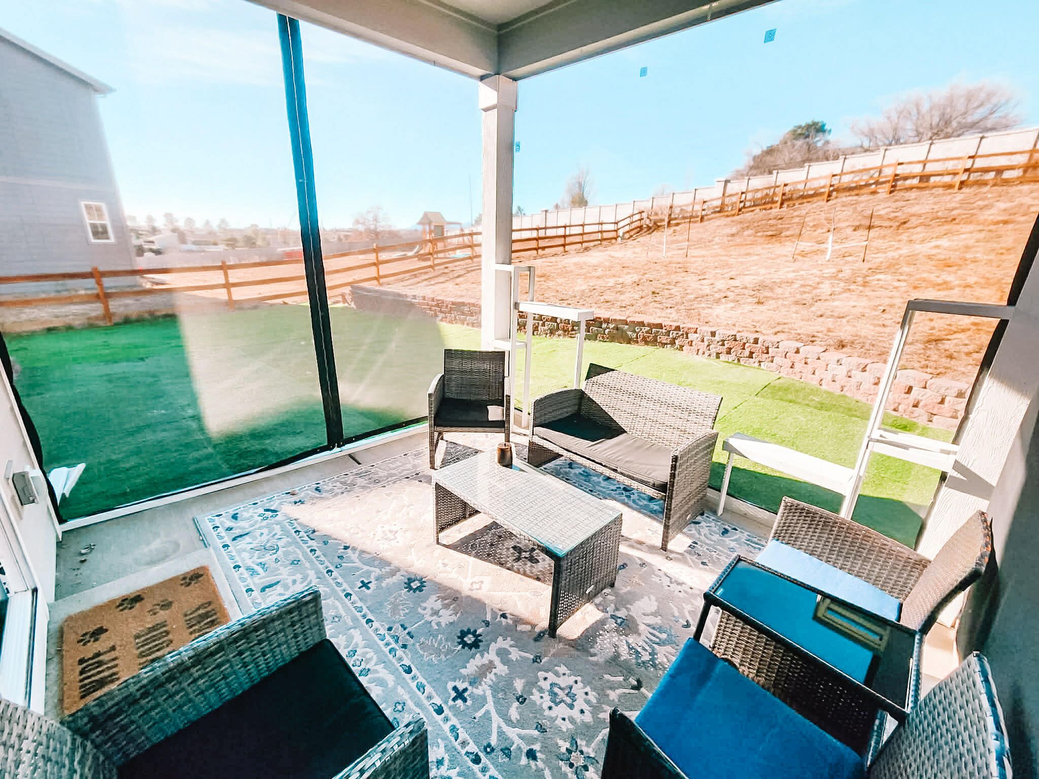 Eine überdachte Terrasse mit Korbmöbeln, einem gemusterten Teppich und Blick auf einen umzäunten Garten.