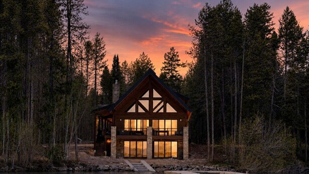 Eine Lodge mit großem Fensterfront, Steinunterbau und Holzfassade, umgeben von Wald und einem Gewässer.