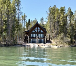A cabin with a large windowed front, surrounded by tall trees and a body of water. - Stillwater Retreat | Waterfront + Private Dock (Island Park)