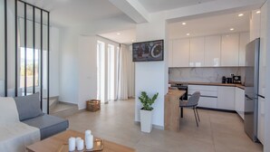 Un salon moderne avec un canapé, une table basse ornée de bougies, un coin cuisine doté de meubles blancs et un grand téléviseur fixé au mur.