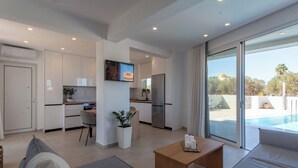 Un salon moderne avec un canapé, une table basse et un coin repas. La cuisine est équipée de meubles blancs et d’un réfrigérateur. Une grande fenêtre offre une vue sur une piscine et des palmiers.
