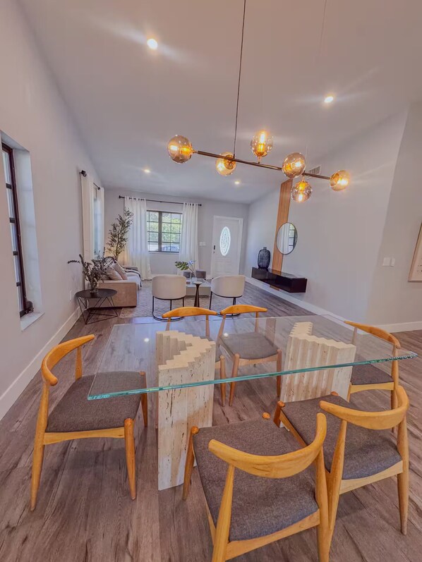 A modern living room with a glass-top coffee table surrounded by wooden chairs, a wooden sideboard, and a chandelier with multiple bulbs. - Enjoyable 3-bedroom house with AC in vibrant Miami (Miami)