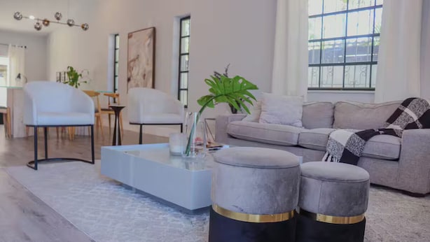 Ein modernes Wohnzimmer mit einem grauen Sofa, weißen Sesseln, einem Glastisch und zwei grauen Poufs mit goldenen Akzenten.