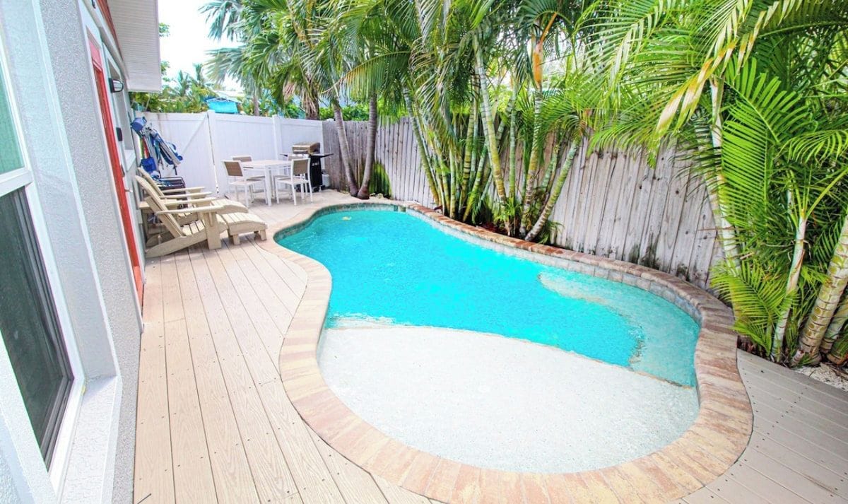 A swimming pool with a wooden deck and lounge chairs, surrounded by a wooden fence and palm trees.