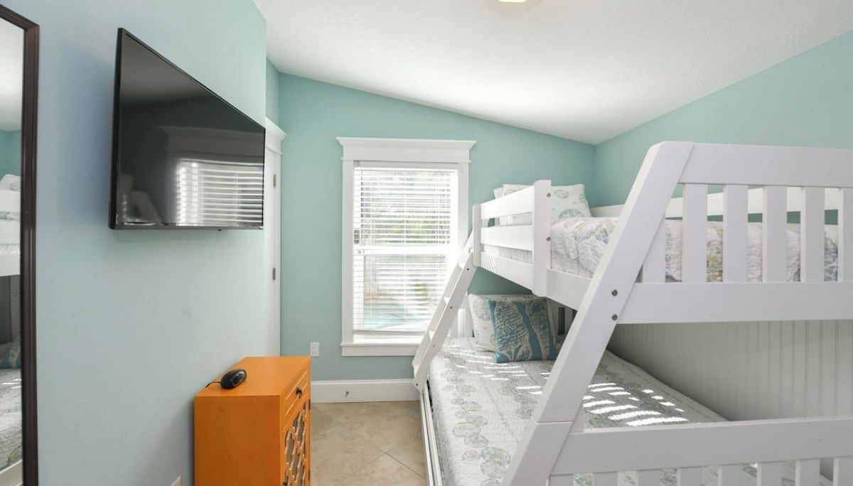 A child's bedroom with bunk beds, a TV, a window with blinds, and a small dresser.