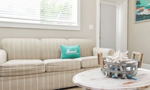 A beige-striped sofa with a teal pillow, a round table with a decorative item, and a window with blinds. - Coconut cottage unit 2! (Holmes Beach)