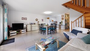 Ein Wohnzimmer mit Fernseher, einem Glastisch, einer Couch, einem Essbereich mit Stühlen und einer Treppe.