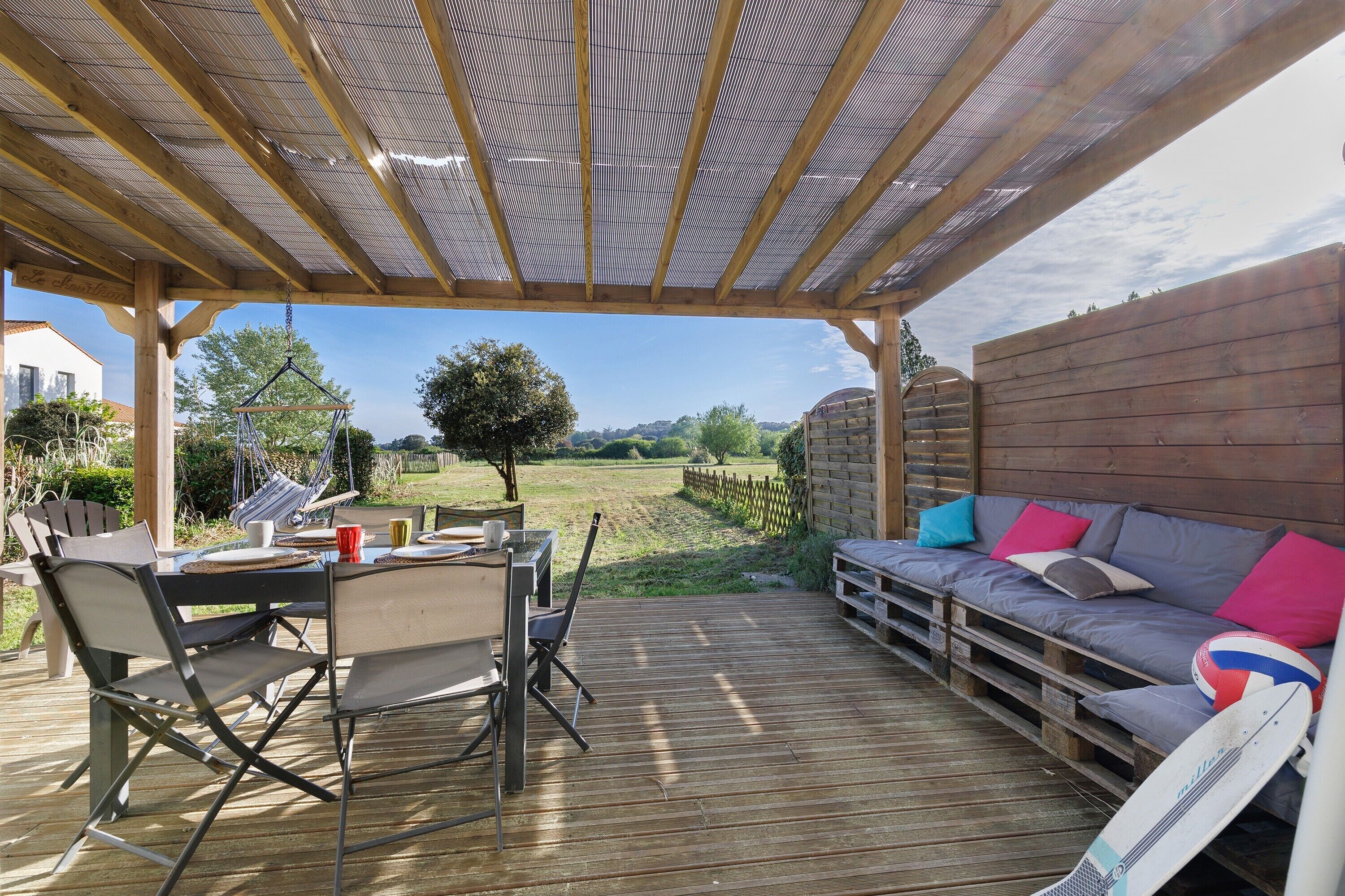 Une terrasse en bois dotée d’un espace couvert, une table à manger avec des chaises et un canapé avec des coussins.
