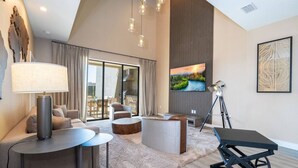 Una sala de estar moderna con sofá, mesa de centro y comedor con sillas. Se aprecia un ventanal con cortinas, un cuadro en la pared y una lámpara sobre una mesa.