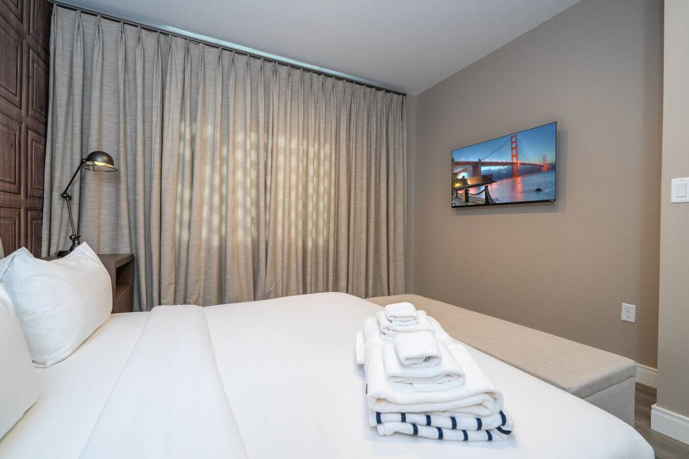 Habitación de hotel con una cama grande, una mesita de noche con lámpara, un cuadro de un puente y toallas dobladas con cuidado sobre la cama.