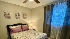 Une chambre à coucher avec un lit, un ventilateur de plafond, une table de chevet, une lampe et une fenêtre avec des rideaux.