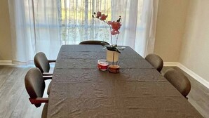 Un coin repas avec une longue table, six chaises, un vase de fleurs et une fenêtre ornée de rideaux légers.