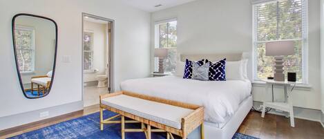 Une chambre à coucher avec un grand lit, un banc, un miroir et deux fenêtres équipées de stores.