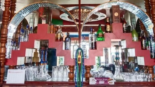 A bar area with a central chandelier, wooden columns, and a variety of bottles and glasses on shelves. - Hotel peace and happy (Bhimeshwar)