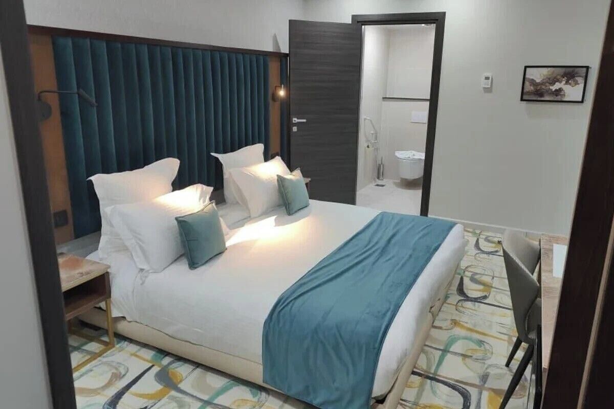 Habitación de hotel con cama, mesitas de noche, una silla, baño con inodoro y lavamanos, y una puerta.