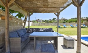 Ein überdachter Außenbereich mit einer Couch, einem Tisch und Stühlen, mit Blick auf einen Pool und eine Steinmauer.