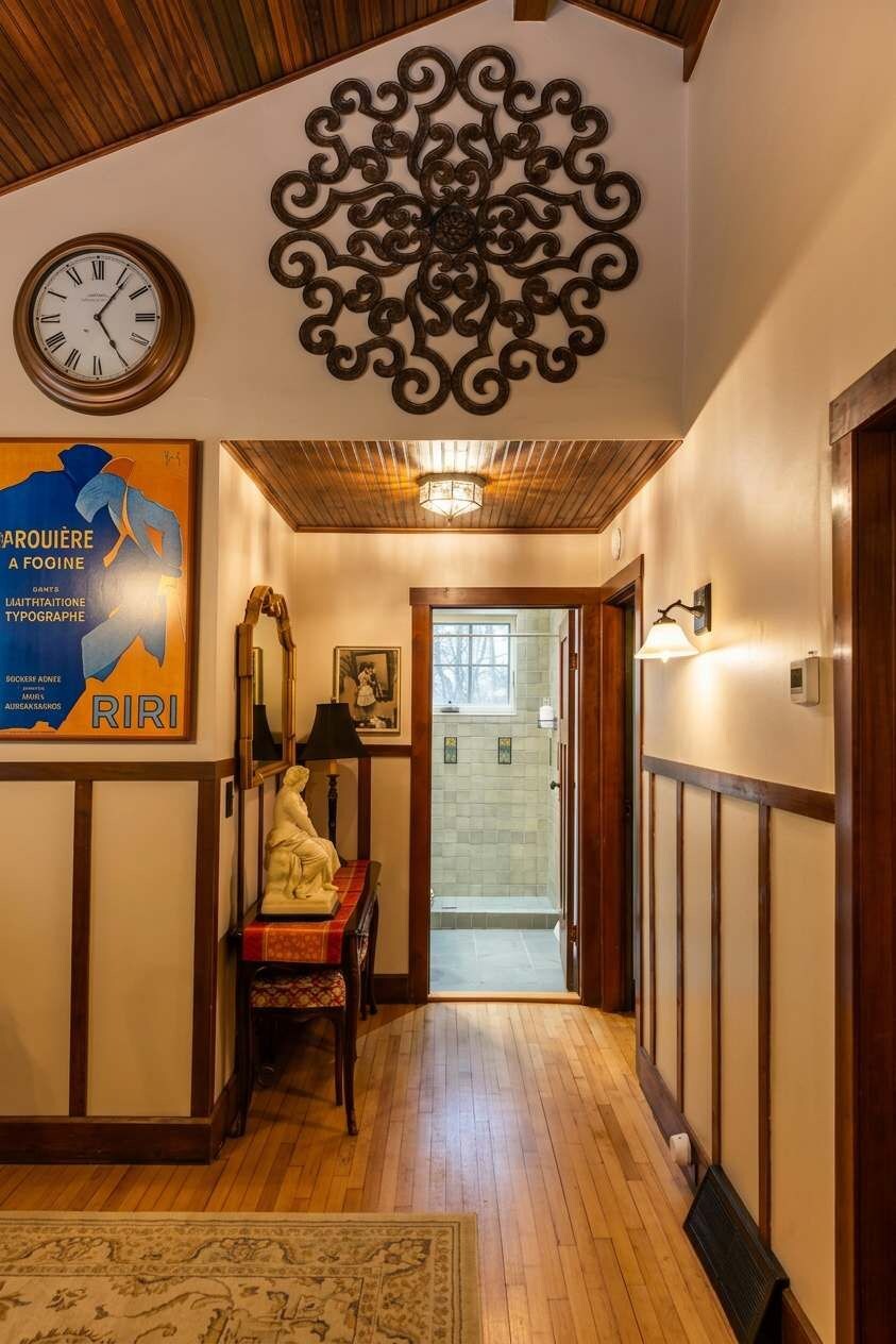 A hallway with a wooden ceiling, a decorative metal piece, a clock, a poster, a table with a lamp, and a door leading to another room.