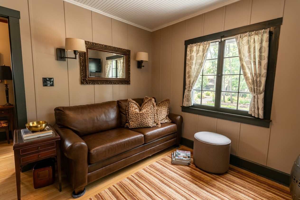 Une pièce comprenant un canapé en cuir marron, un pouf rond, une table d’appoint en bois avec un bol et des livres, un miroir avec des appliques murales et une fenêtre ornée de rideaux à motifs.