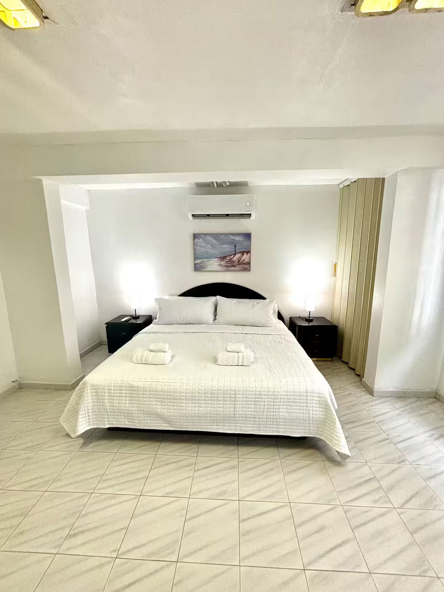 Un dormitorio con cama, dos mesitas de noche, aire acondicionado de pared, un cuadro y una ventana con cortinas.