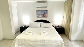 Une chambre à coucher comprenant un lit, deux tables de chevet, un climatiseur fixé au mur, un tableau et une fenêtre avec des rideaux.