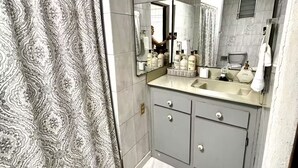 Une salle de bain avec un rideau de douche à motifs, un grand miroir, un lavabo avec des articles de toilette et des meubles gris.