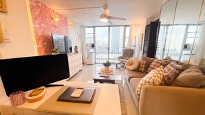 Un soggiorno moderno con una TV a schermo piatto, una scrivania con un portatile, un divano con cuscini decorati e un balcone con vista sulla città.