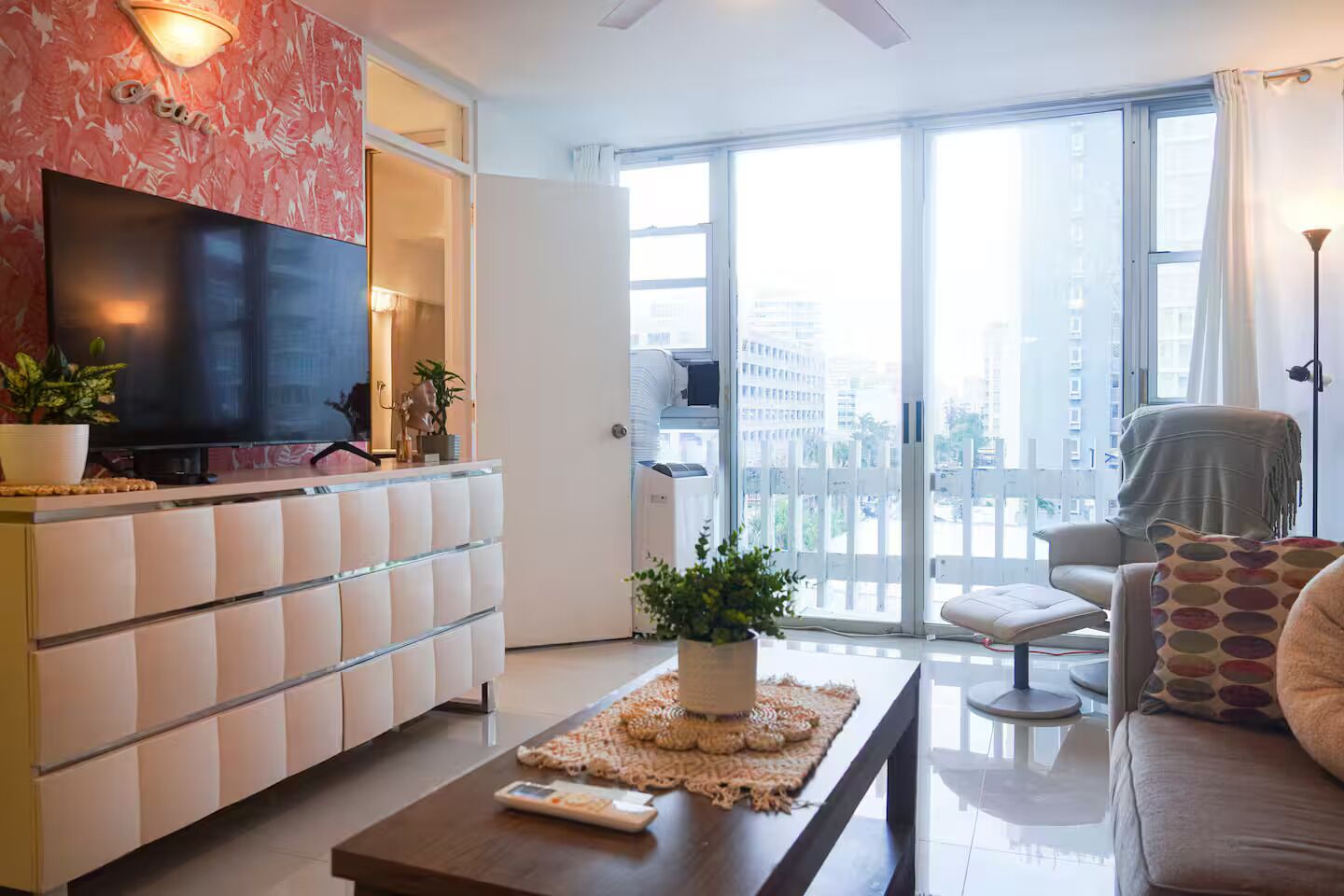 A modern living room with a flat-screen TV, a sofa, a coffee table, and a balcony with a view of the city.
