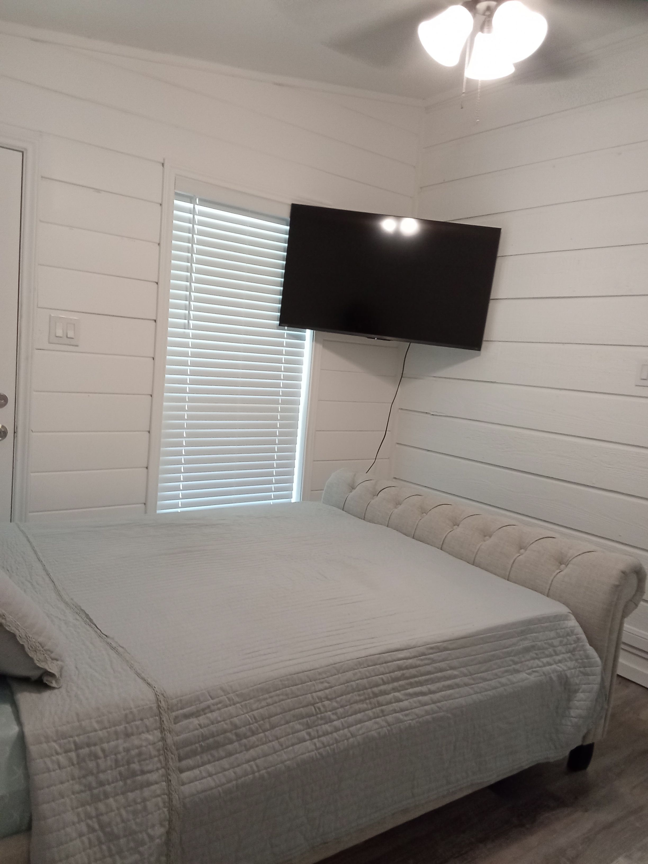 A bedroom with a bed, a television mounted on the wall, and a window with blinds.