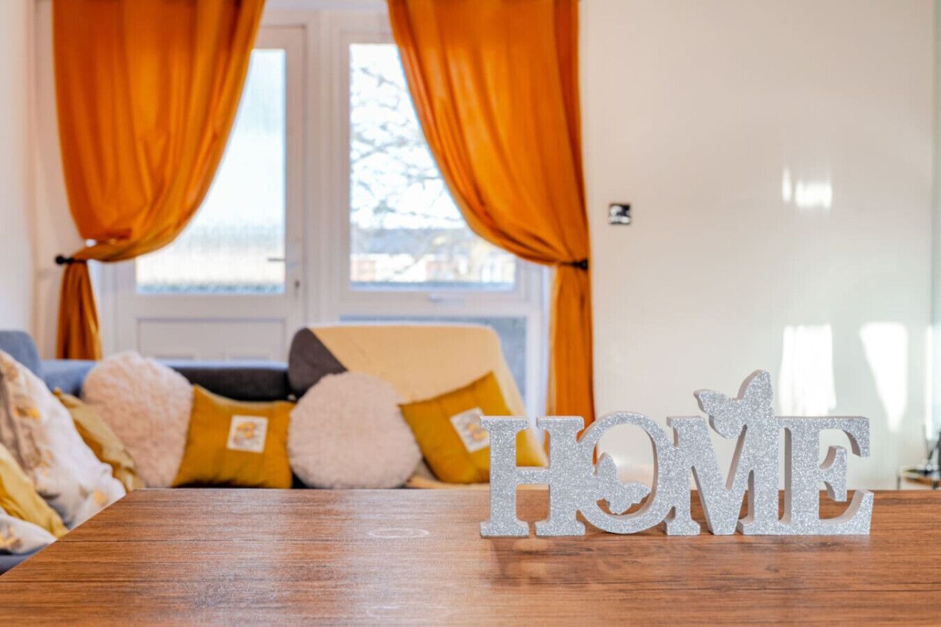 A room with a wooden table displaying the word "HOME" in glittery letters, a sofa with cushions, and orange curtains.