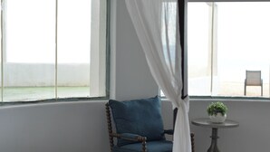 Ein Schlafzimmer mit Himmelbett, einem Sessel mit blauem Kissen, einem kleinen runden Tisch mit Pflanze und Meerblick durch große Fenster.