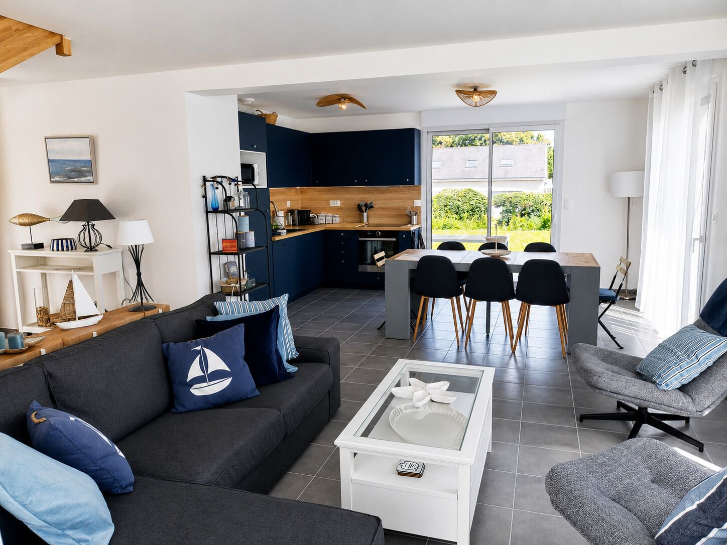 A modern living room with a sofa, armchair, and coffee table, a kitchen with a dining area, and a large window offering a view of greenery.