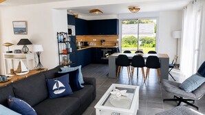 Un salon moderne comprenant un canapé, un fauteuil et une table basse, une cuisine avec un coin repas, et une grande fenêtre donnant sur un paysage verdoyant.