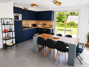 A modern kitchen with dark blue cabinets, a wooden dining table, and a view of the garden. - “L'Escale Bleue” - on the Rhuys Peninsula, close to unspoiled nature (Le Tour-du-Parc)