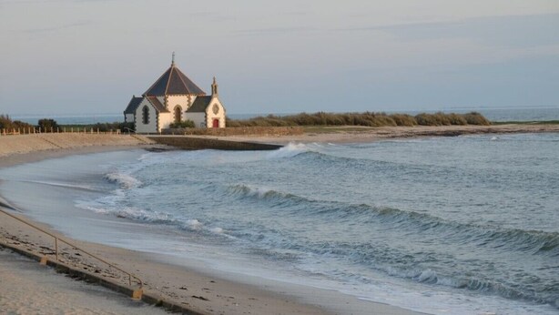 Un bâtiment situé sur la côte, doté d’un toit en pente et d’une petite tour, et qui se trouve à proximité d’une plage de sable où les vagues sont douces.