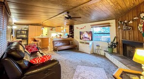 A cozy living room with a leather sofa, a fireplace, and a television. There is a kitchen area in the background with wooden cabinets and a dining table. - Ruidoso Cabin: Mountain Views & Private Deck (Ruidoso)
