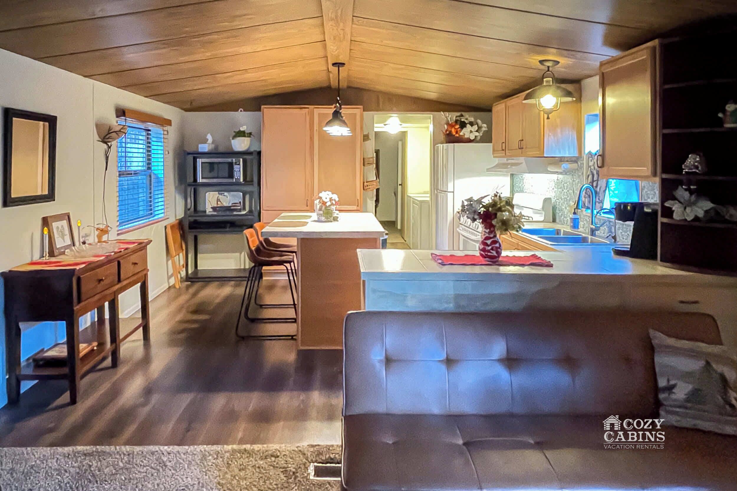 Interior acogedor de una cabaña con techo de madera, un comedor con mesa y sillas, una cocina con microondas y fregadero, y una sala de estar con sofá.
